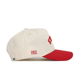 The Upside Down Scheels Hat is a cream baseball cap with a red brim, made from high-quality cotton. It features "1902" embroidered in red on the side, partial red lettering on the front, a small white tag at the back, and an adjustable snap closure.