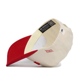 The Upside Down Scheels Hat is a cream-colored snapback with a red brim, shown inside-out to reveal the NO RIVALS label and QR code. "1982" is embroidered in red on the side of this high quality cotton cap.