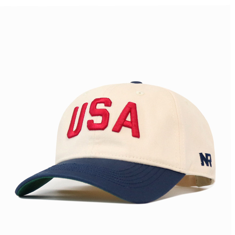 Off-white, unstructured USA Vintage Hat - 6 Panel Unstructured with a navy blue brim, featuring red USA lettering on the front and a small navy No Rivals logo on the side.