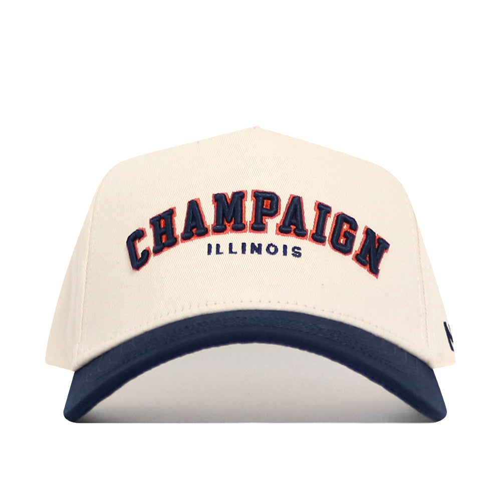 The No Rivals Champaign Hometown Hat features a cream, structured snapback design with "CHAMPAIGN" in bold navy letters outlined in orange, "ILLINOIS" below in blue, and a soft navy blue cotton curved brim.