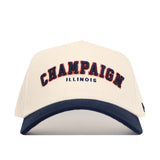 The No Rivals Champaign Hometown Hat features a cream, structured snapback design with "CHAMPAIGN" in bold navy letters outlined in orange, "ILLINOIS" below in blue, and a soft navy blue cotton curved brim.