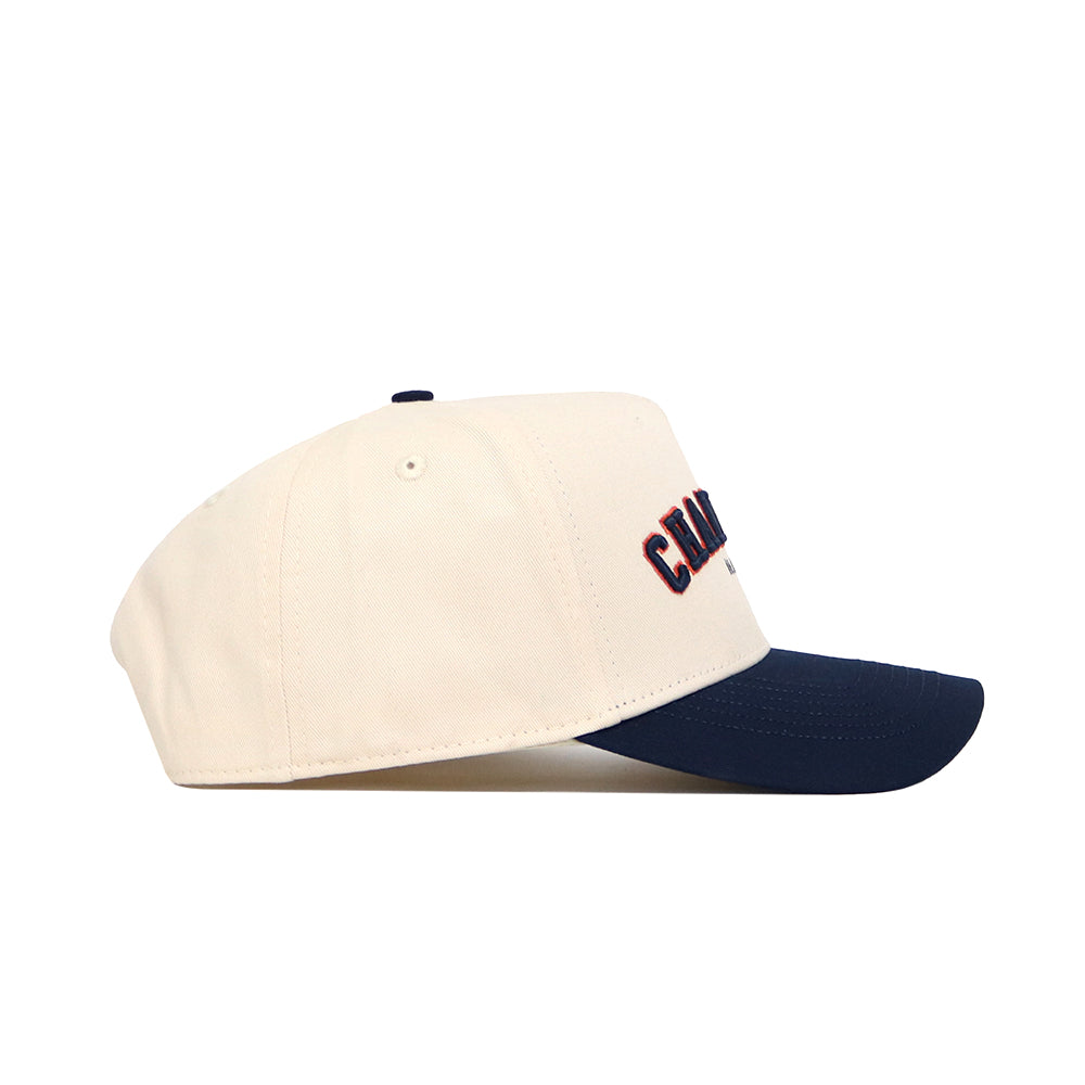 The No Rivals Champaign Hometown Hat is a cream cotton baseball cap with a navy curved brim, seen from the side. The structured fit shows partially visible navy and orange embroidery on the front.