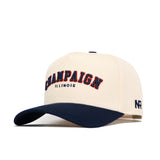 The No Rivals Champaign Hometown Hat is a cream cotton snapback cap with a navy brim, featuring "CHAMPAIGN" in large navy and red letters above "ILLINOIS" in smaller navy text on the structured front.