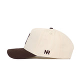 The No Rivals University of Illinois Espresso Hat is a cream cotton baseball cap with a dark brown brim, seen from the side. "NR" is embroidered in brown thread on the side.