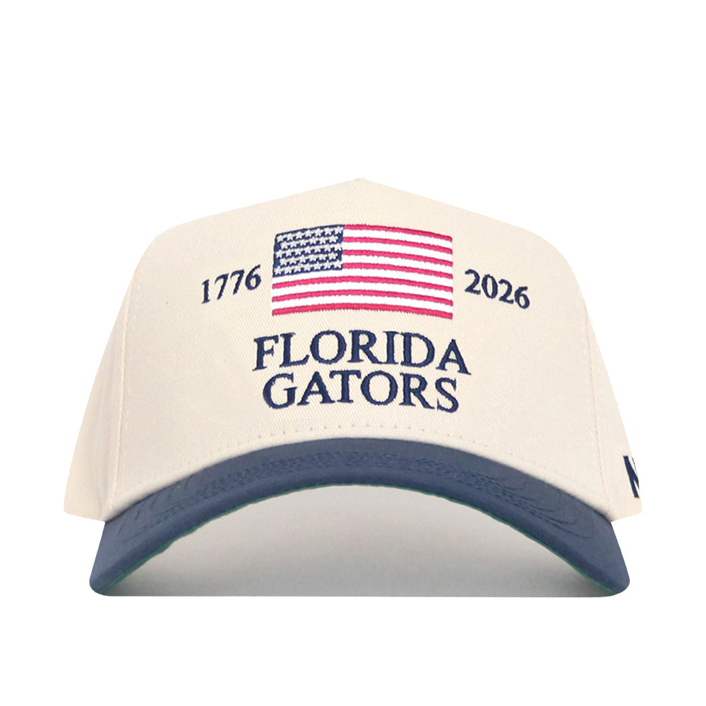 The No Rivals Florida Gators 250th Americana Hat is a cream snapback with an official US flag, embroidered FLORIDA GATORS logo, “1776” and “2026” flanking the flag, and a dark blue brim.