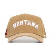 The Montana Rustic Hat by No Rivals is a tan cotton canvas snapback with a structured fit, featuring MONTANA in large white letters on the front and a red bear paw symbol on the left side.