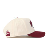The No Rivals Montana Grizzlies Stadium Hat is a cream-colored snapback baseball cap featuring a maroon brim, an officially licensed maroon bear paw print embroidered on the side, and a partially visible logo on the front. This hat blends classic style with distinctive, embroidered details for a standout look.