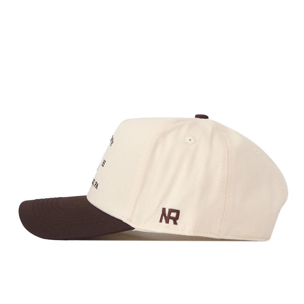 A side view of the Penn State Outlaw Hat - Classic by No Rivals, featuring a cream-colored crown with a brown brim. Near the back, an embroidered “NR” logo in brown thread adds detail, while its classic gameday fit ensures ultimate comfort.