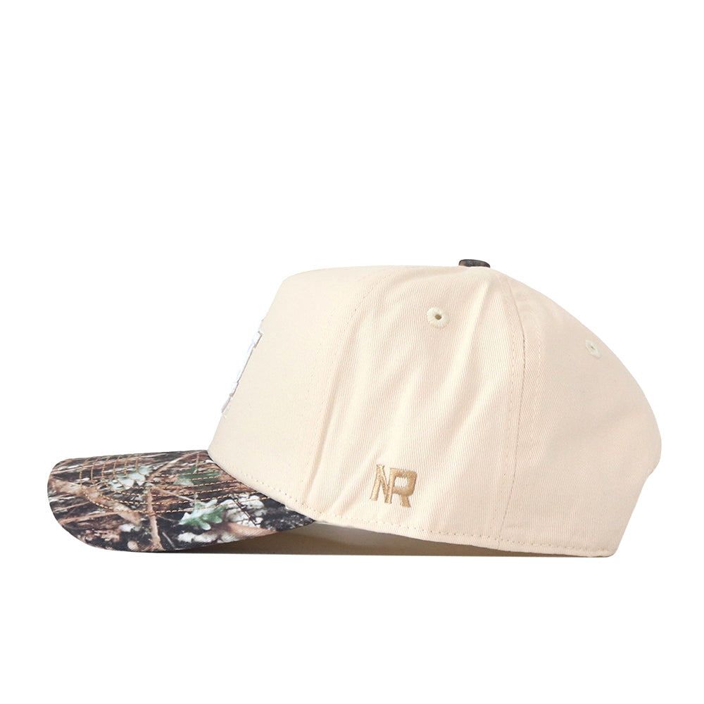A side view of the Auburn Camo Connector Hat by No Rivals, featuring a beige crown with a camouflage-patterned brim and accented by a small gold NR embroidered logo near the back—an effortlessly stylish addition to your gameday look.