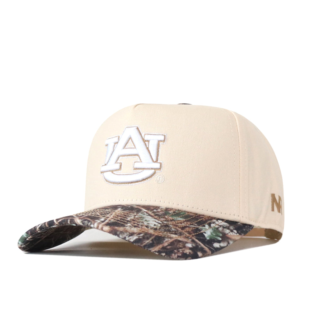 Show off your gameday style with the Auburn Camo Connector Hat by No Rivals. This beige snapback features a white AU embroidered logo on the front and a camo-patterned brim, while a small gold emblem accents the side for an extra touch of flair. The hat is displayed at an angle against a white background.