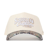 The Scheels Camo Connector Hat is a beige snapback baseball cap made of high-quality cotton, featuring "Scheels Outfitters Since 1902" embroidered in white and brown with two tree graphics and a camouflage-patterned bill.