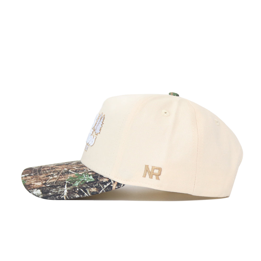 The Scheels Camo Connector Hat features a beige crown, camo-patterned brim, high-quality cotton build, embroidered white front detail, and gold NR initials on the side. A snapback closure provides a comfortable fit. Shown on a white background.