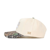 The Scheels Camo Connector Hat features a beige crown, camo-patterned brim, high-quality cotton build, embroidered white front detail, and gold NR initials on the side. A snapback closure provides a comfortable fit. Shown on a white background.