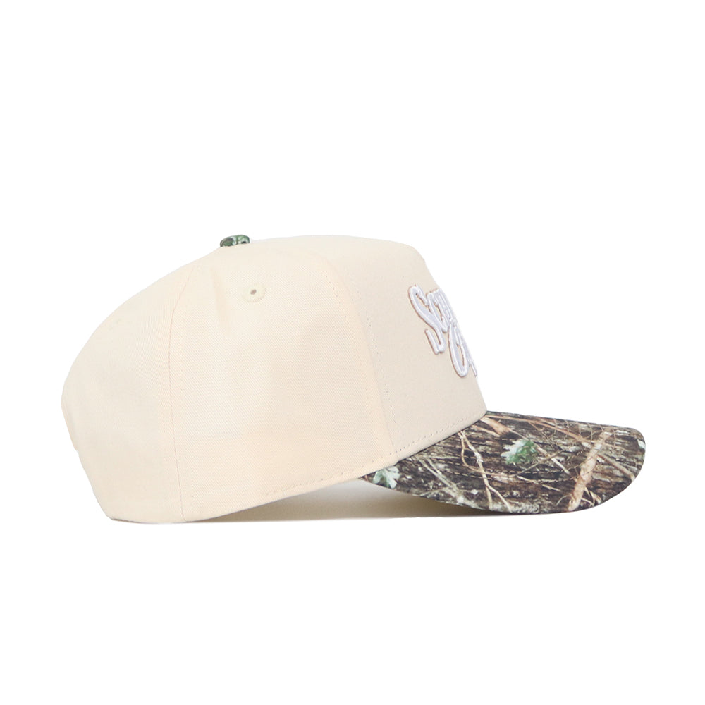 The Scheels Camo Connector Hat is a beige, high-quality cotton baseball cap with a camouflage-patterned brim and white front embroidery, displayed side-on against a white background.