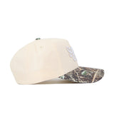 The Scheels Camo Connector Hat is a beige, high-quality cotton baseball cap with a camouflage-patterned brim and white front embroidery, displayed side-on against a white background.