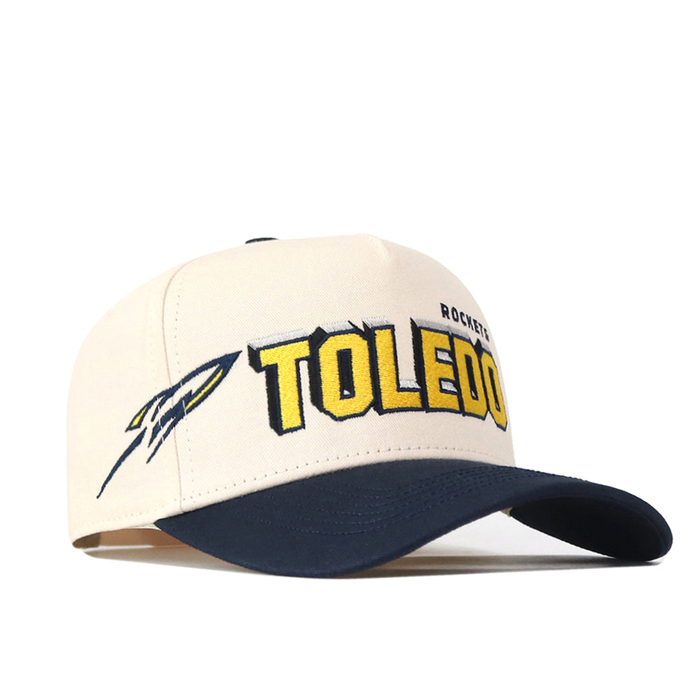 The No Rivals Toledo Alma Mater Hat features a beige and navy design with “TOLEDO” in yellow letters, the officially licensed Rocket logo, and partial “ROCKETS” text embroidered on the front. It includes an adjustable snap back for a comfortable fit.