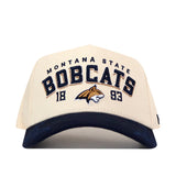 The No Rivals Montana State Rally Hat is a beige snapback featuring a blue brim. “Montana State Bobcats” and “1893” are embroidered on the front, along with an officially licensed bobcat logo at the center.