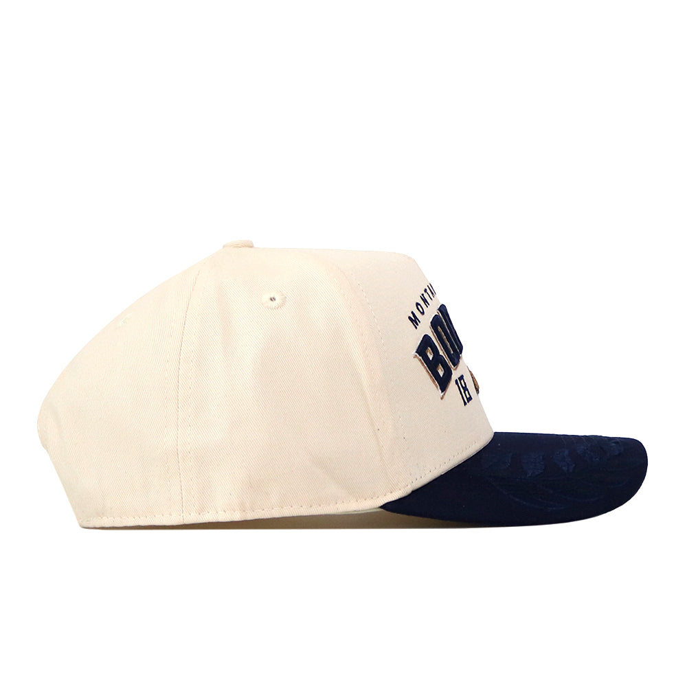 A No Rivals Montana State Rally Hat in cream with a dark blue brim, shown from the left side. The front panel features an officially licensed, embroidered logo and partially visible text.
