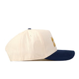 A side view of the No Rivals WVU Mountaineers Script Overlay Mascot Patch Hat, a beige snapback baseball cap with a navy blue brim. The front displays an officially licensed yellow embroidered Mountaineers logo, and the side panel includes a small ventilation hole.