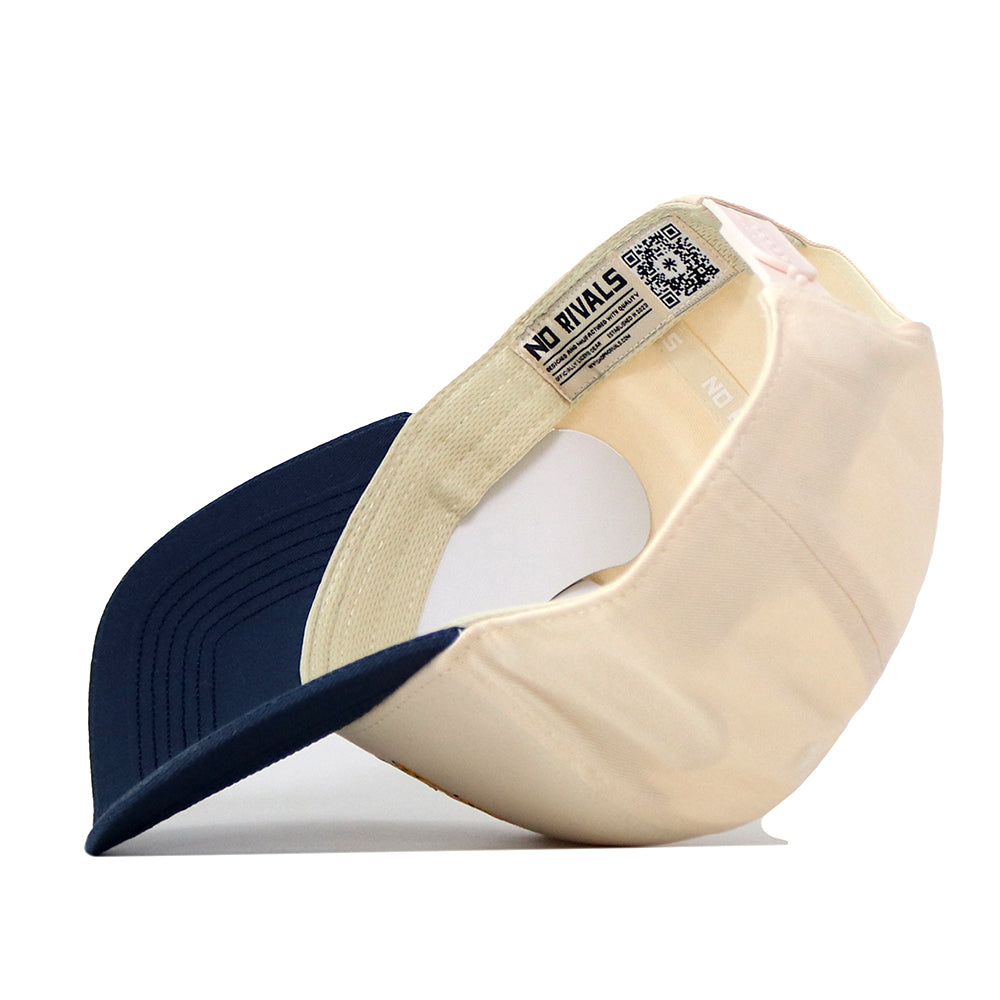 The WVU Mountaineers Script Overlay Mascot Patch Hat by No Rivals is pictured upside down, revealing the interior label featuring the NO RIVALS branding and a QR code. The hat boasts a navy brim paired with a beige crown, and its classic snap back closure offers an adjustable fit.