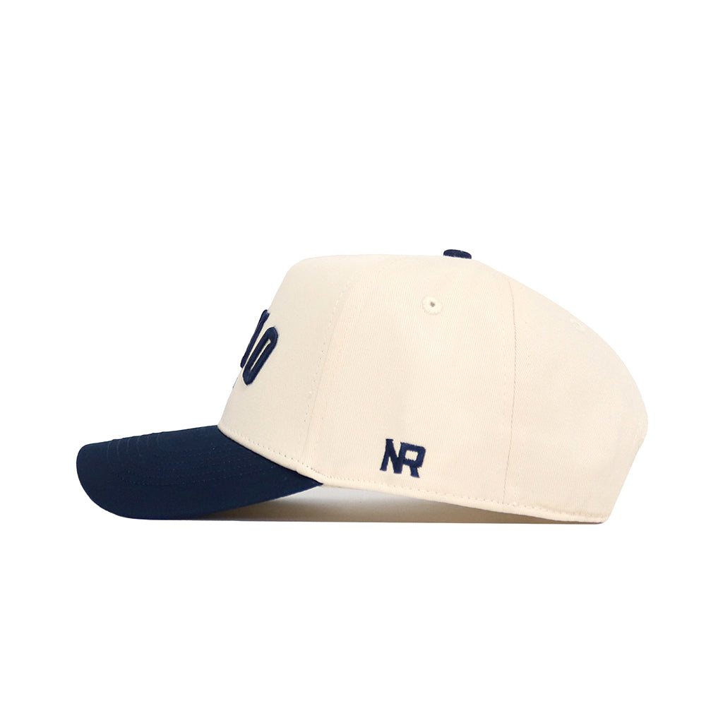 The Reno Hometown Hat by No Rivals is a cream-colored cotton baseball cap with a navy blue brim, shown in side view. It has a structured fit and features dark blue embroidered "NR" letters on the side.