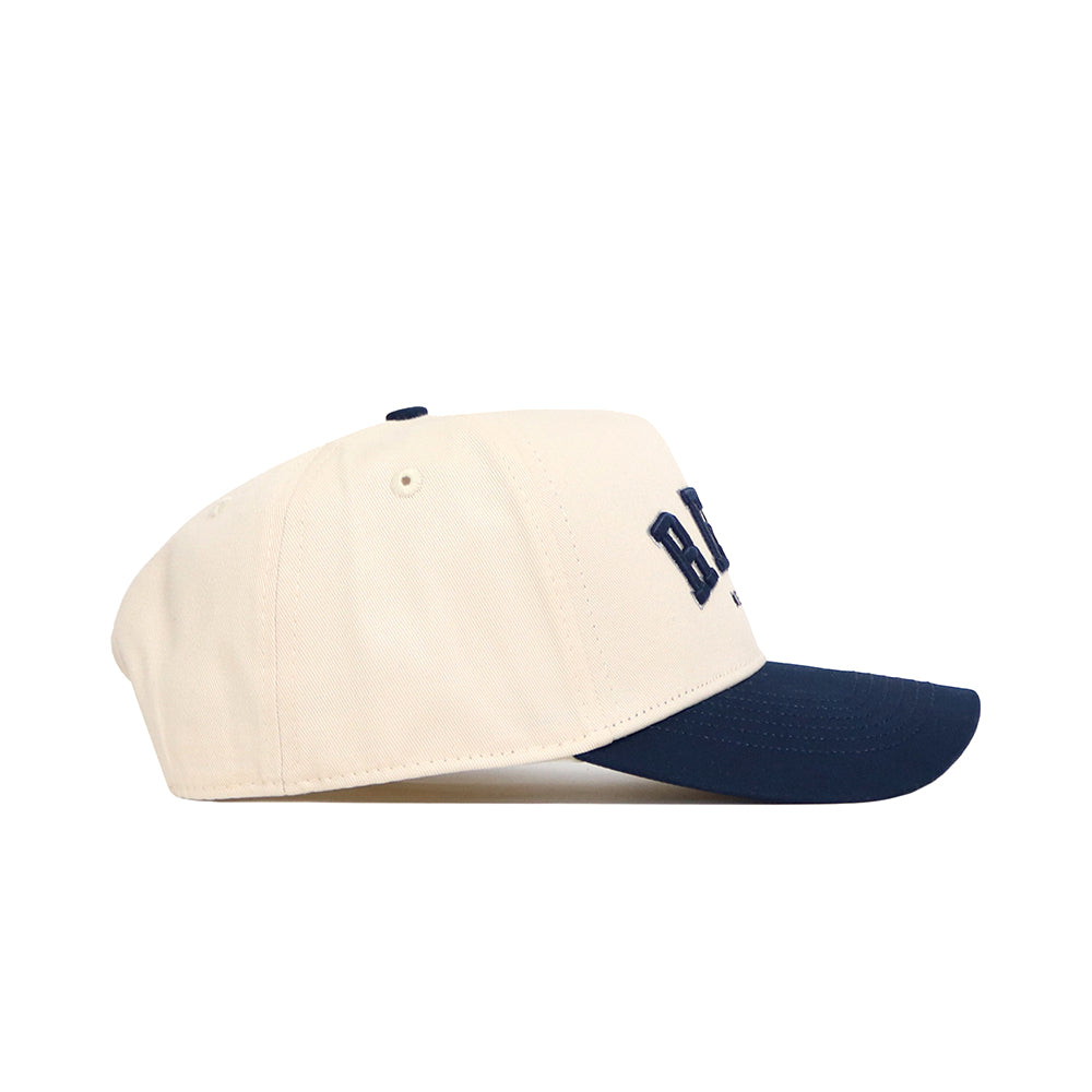 The No Rivals Reno Hometown Hat is a beige cotton baseball cap with a navy curved brim and partially visible dark blue embroidery on the front, pictured from the side against a white background.