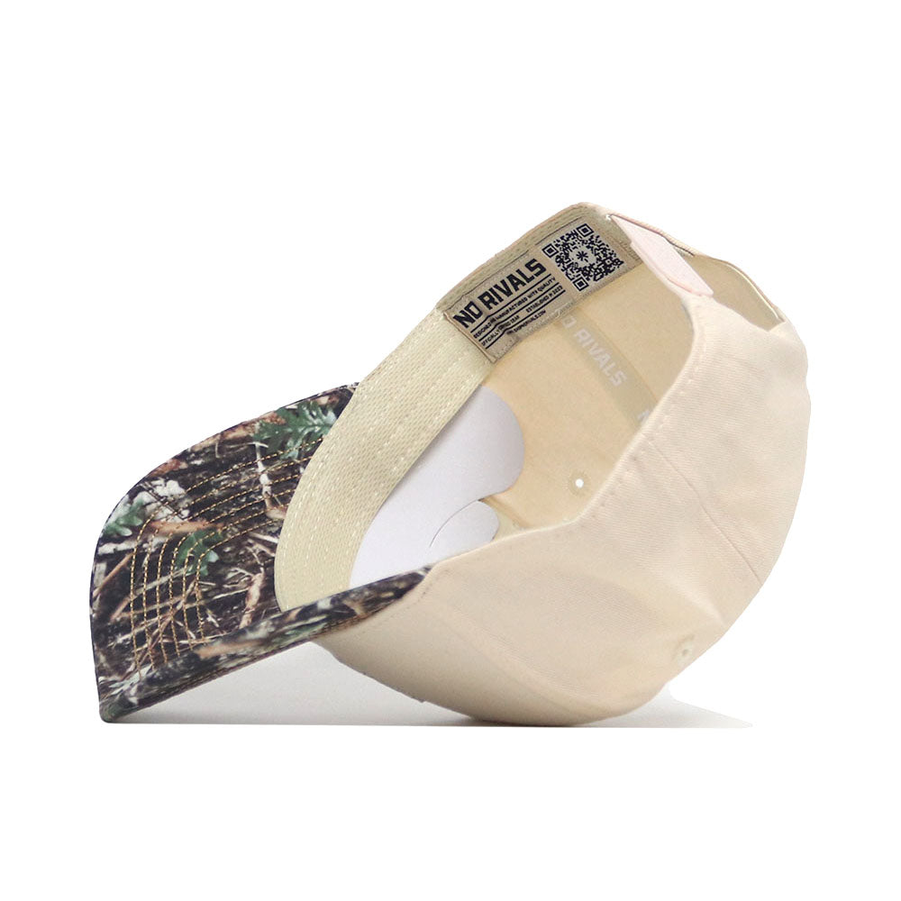 The Kansas Camo Connector Hat by No Rivals is pictured upside down, showing its camo-patterned brim and interior "NO RIVALS" label. Made from premium cotton, the beige snap back cap rests on a white surface.