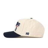 Introducing the Duquesne Stride Hat by No Rivals—a beige baseball cap with a navy brim, adorned with embroidered logos, including side embroidery that reads “NR,” and front text in striking blue and red. The snap back closure ensures an adjustable, comfortable fit.
