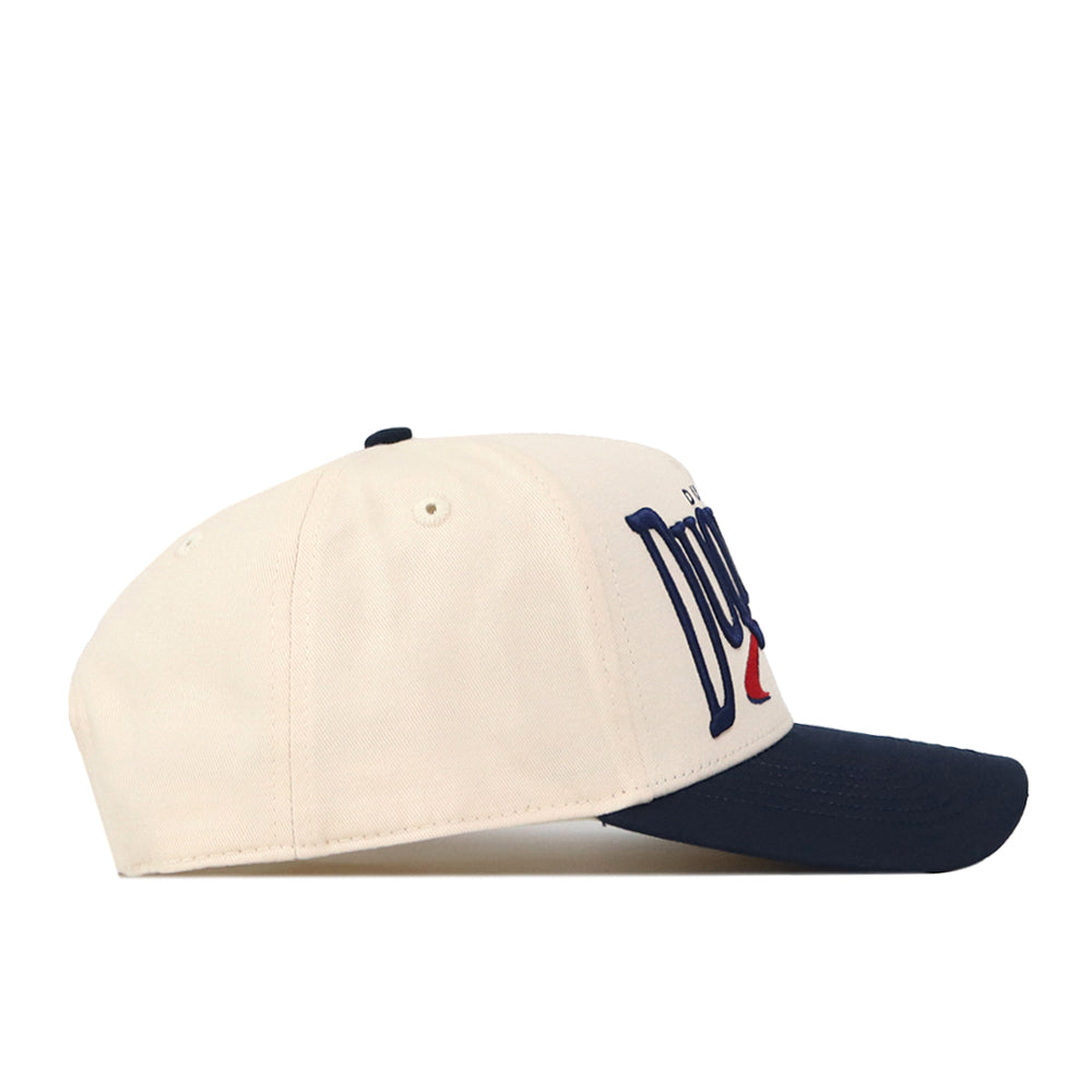 The Duquesne Stride Hat by No Rivals is an officially licensed, cream-colored baseball cap with a navy blue brim, displayed from the side. It features partially embroidered blue and red text on the front panel and has a classic snapback closure for an adjustable fit.