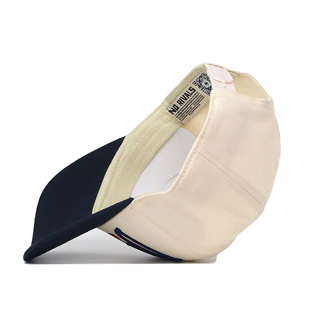 A cream and navy blue Duquesne Stride Hat by No Rivals is propped on its side, displaying the inside label, inner stitching, and adjustable snapback strap at the back.