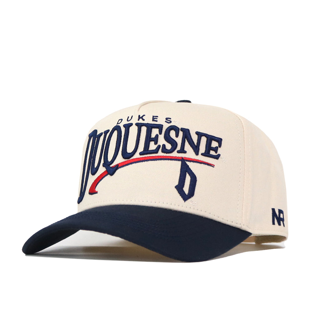 Introducing the Duquesne Stride Hat by No Rivals: a beige and navy baseball cap with bold, embroidered “DUQUESNE” lettering, “DUKES” above, and a blue “D” with red swoosh detail underneath. This cap features a curved brim, an “NR” logo on the side, and an adjustable snap back for a comfortable fit.