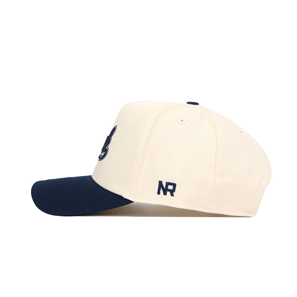 The No Rivals Nevada Logo Patch Hat is a cream snapback baseball cap with a dark blue brim and an official 3D NR logo embroidered on the side, displayed in a left side profile against a white background.