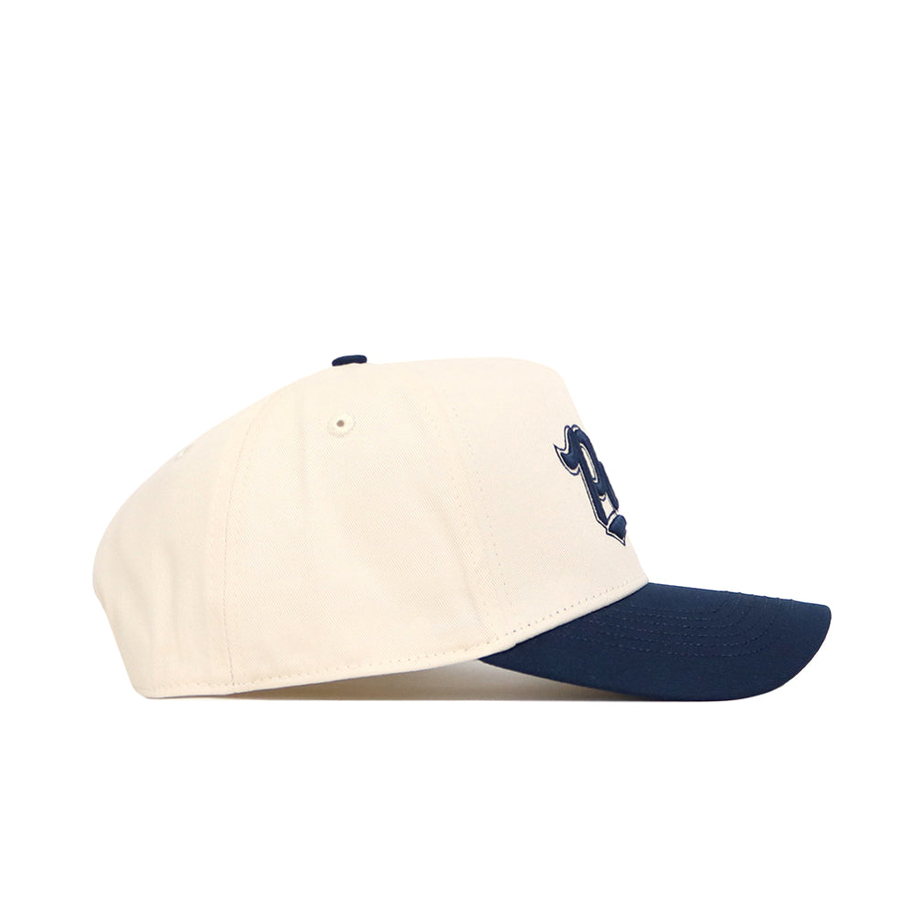 The No Rivals Nevada Logo Patch Hat features a cream crown, navy blue brim, and a 3D embroidered stylized D logo on the front, displayed in a side profile view against a white background.