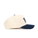 The No Rivals Nevada Logo Patch Hat features a cream crown, navy blue brim, and a 3D embroidered stylized D logo on the front, displayed in a side profile view against a white background.