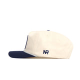 Side view of the Penn State Seal Hat by No Rivals, a cream snapback with a navy brim and NR embroidered in navy on the side, crafted from high-quality cotton.