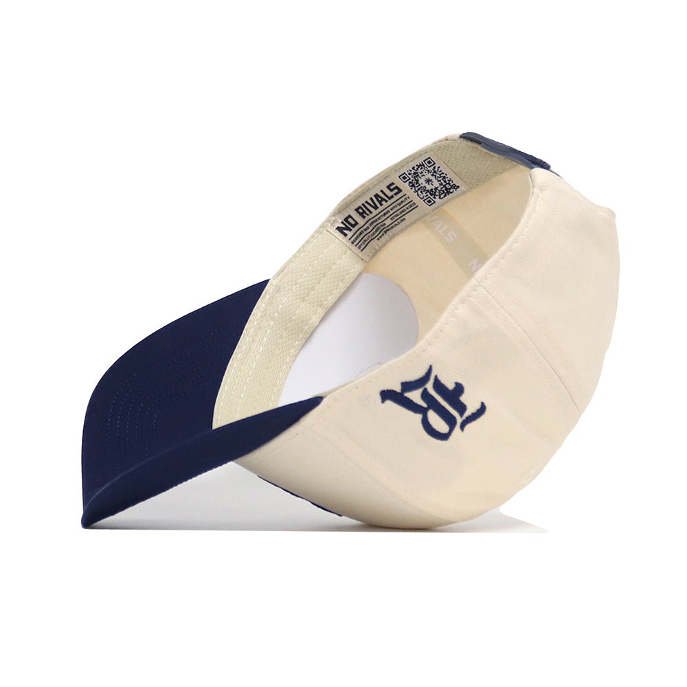 The Rice Sammy Sailor Patch Hat by No Rivals is a cream and navy blue snapback cap. Shown upside down, it features a navy underside brim, an inside "NO RIVALS" tag, embroidered patch logo, and an adjustable closure for comfort.