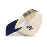 The Rice Sammy Sailor Patch Hat by No Rivals is a cream and navy blue snapback cap. Shown upside down, it features a navy underside brim, an inside "NO RIVALS" tag, embroidered patch logo, and an adjustable closure for comfort.