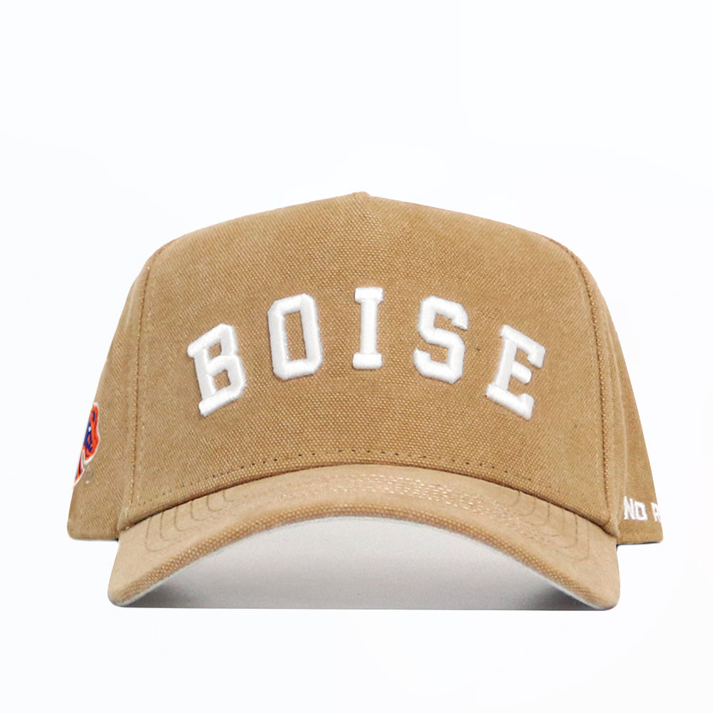 The Boise State Rustic Hat by No Rivals is a tan cotton canvas baseball cap with a structured fit, large white "BOISE" embroidery on the front, and a small colorful patch on the left side.