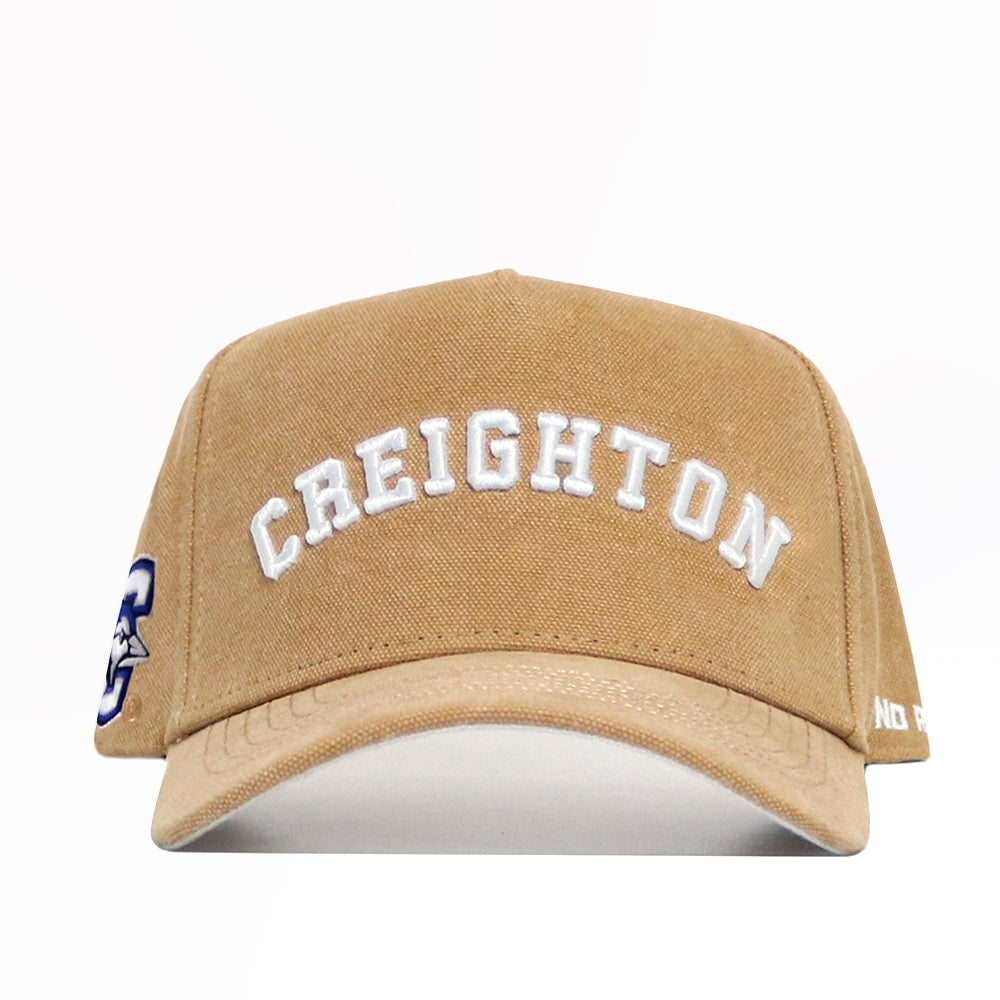 The Creighton Rustic Hat by No Rivals is a tan cotton canvas snapback cap featuring “CREIGHTON” embroidered in white on the front, side logo, partial white text on the brim, and is officially licensed.