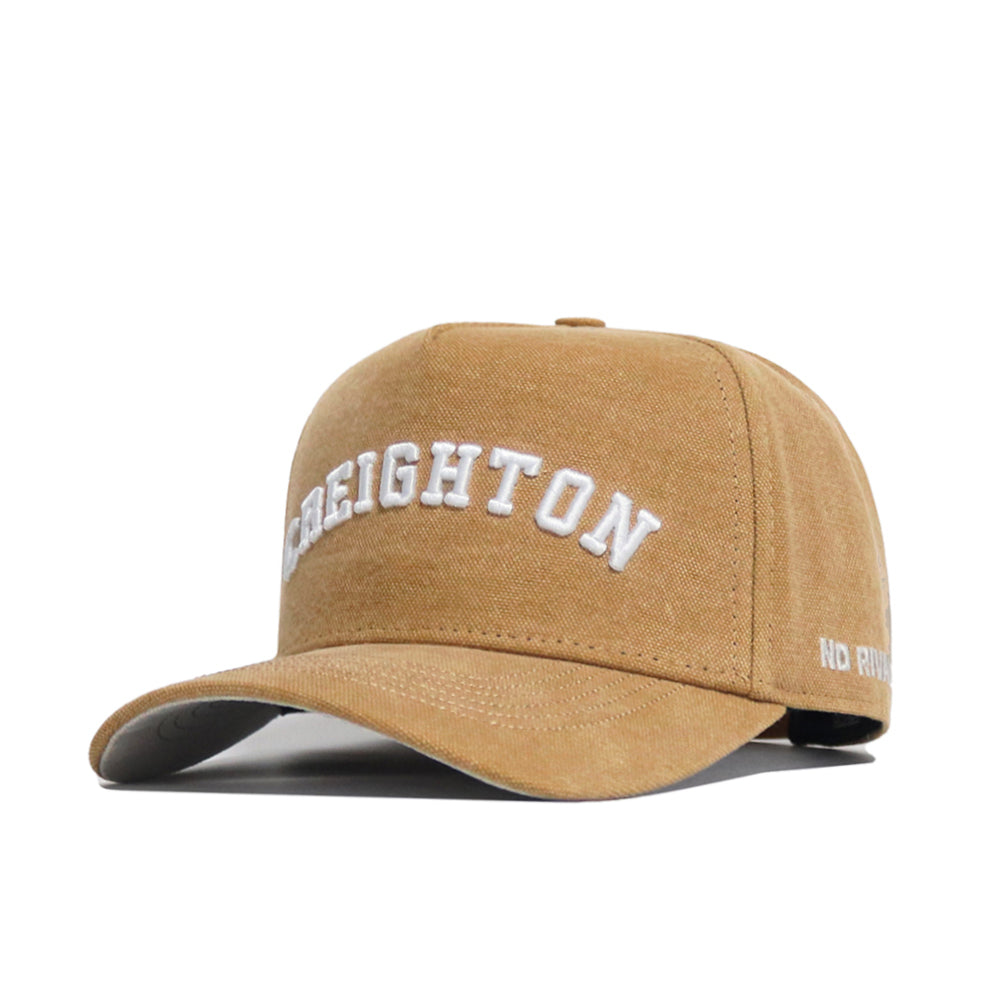 The Creighton Rustic Hat by No Rivals is a tan cotton canvas cap with "CREIGHTON" embroidered in white on the front, "NO RIVALS" on the side, a classic snapback closure, and is officially licensed.