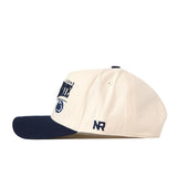 Introducing the Penn State Nittany Lions Stride Hat by No Rivals—a cream-colored baseball cap with a navy blue brim, made from high quality cotton. This structured-fit hat features embroidered text and logos on the front and side, including the letters “NR” stitched on the snapback.