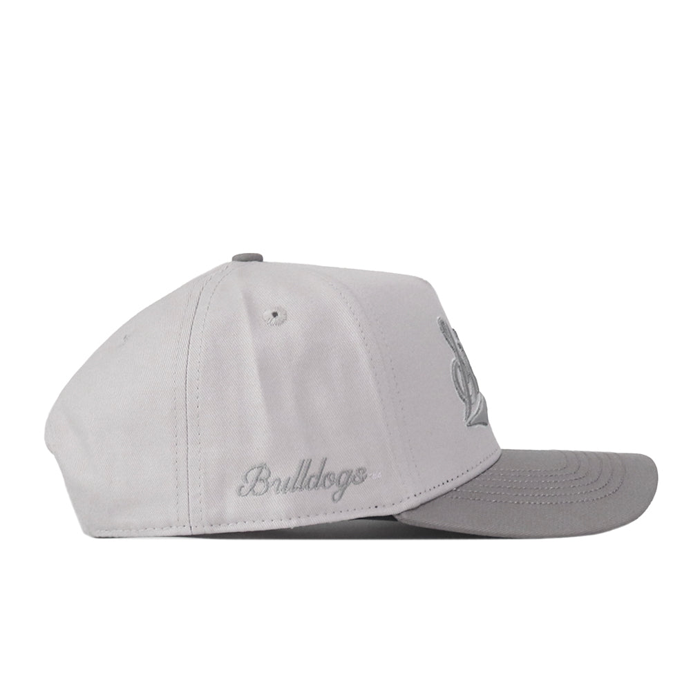 Introducing the No Rivals Mississippi State Graphite Hat: this light gray baseball cap features a flat brim and an adjustable snap back closure for your ideal gameday fit. “Bulldogs” is embroidered in gray script on the side, while a raised embroidered logo appears partially visible on the front panel.