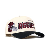 The No Rivals Texas A&M Alma Mater Hat is a beige and black snapback made from premium cotton, with a maroon football helmet and AGGIES embroidered, plus TEXAS A&M above—an officially licensed cap for fans.