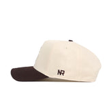 The Marquette Espresso Hat by No Rivals is a cream baseball cap with a dark brown brim, featuring brown "NR" embroidery on the side. This structured snapback is made from premium cotton for comfort and durability.