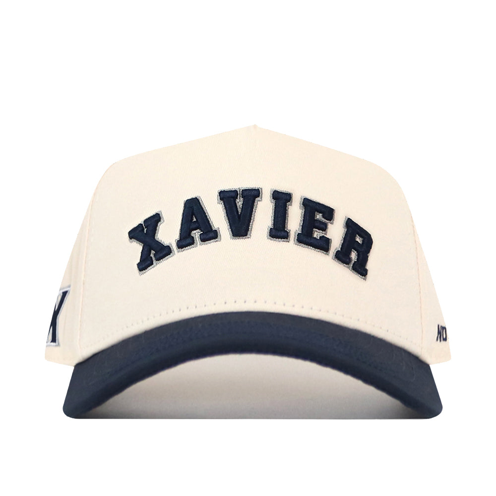 The Right Side Up Xavier Hat - Classic by No Rivals features a beige design with “XAVIER” boldly embroidered in large black letters across the front and a striking black brim. An additional embroidered logo adds to its classic look, making this cap the perfect finishing touch for your gameday outfit. Shown against a plain white background.