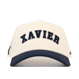 The Right Side Up Xavier Hat - Classic by No Rivals features a beige design with “XAVIER” boldly embroidered in large black letters across the front and a striking black brim. An additional embroidered logo adds to its classic look, making this cap the perfect finishing touch for your gameday outfit. Shown against a plain white background.