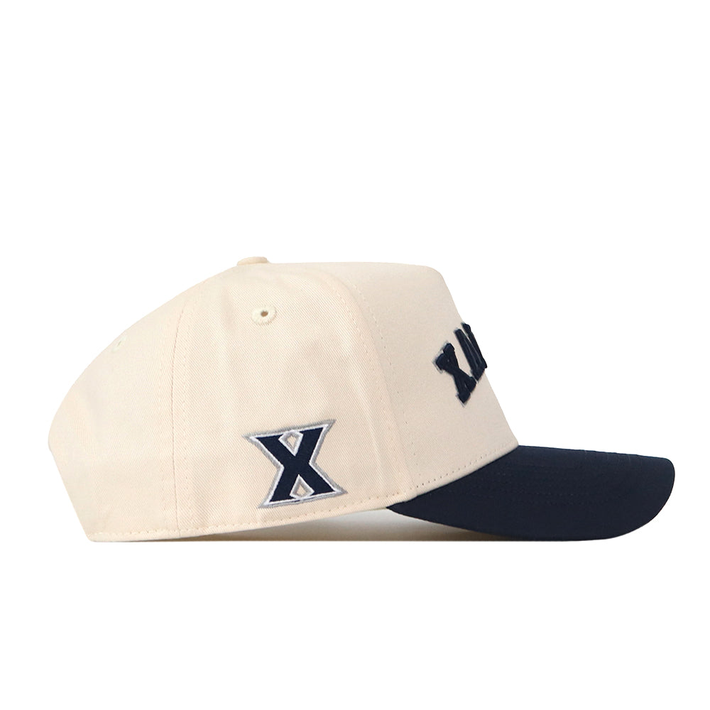 Introducing the No Rivals Right Side Up Xavier Hat - Classic: a cream-colored baseball cap with a navy blue brim. The side displays a dark blue “X” outlined in white, while the front showcases part of a raised, embroidered navy blue logo. A snapback closure ensures the perfect fit for gameday.