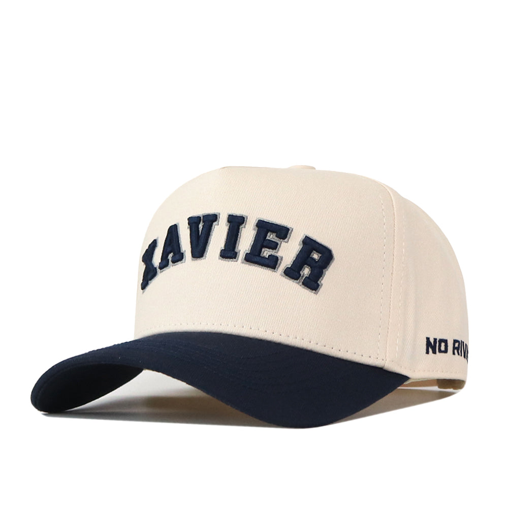 Sure! Here’s the rewritten sentence using your product data: The Right Side Up Xavier Hat - Classic by No Rivals features a beige base with a bold, navy embroidered “XAVIER” logo on the front and a contrasting navy brim. “NO RIVALS” is subtly stitched on the side, offering a classic gameday fit.
