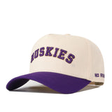 Introducing the Right Side Up Huskies Hat - UW - Classic by No Rivals: This beige baseball cap features a striking purple brim. “HUSKIES” is officially licensed and embroidered in bold purple letters on the front, while “NO RIV” is stitched in purple on the side. The hat includes a snap back adjustment for a comfortable fit.