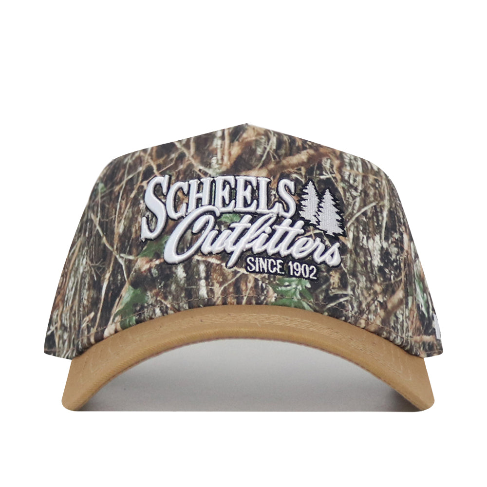 The Scheels Camo Explorer Hat is a camouflage baseball cap with a tan brim, structured fit, snapback closure, white “Scheels Outfitters Since 1902” embroidery, and two tree graphics on the front.